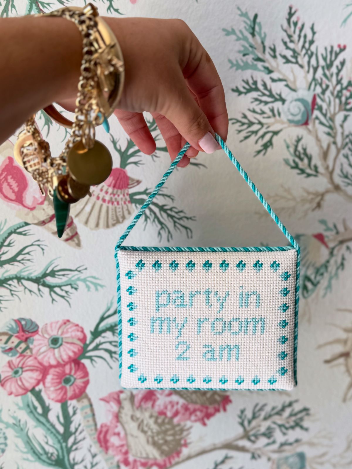 Hand holding a small needlepoint sign with nursery 'party in my room 2 am' text against a seashell background.